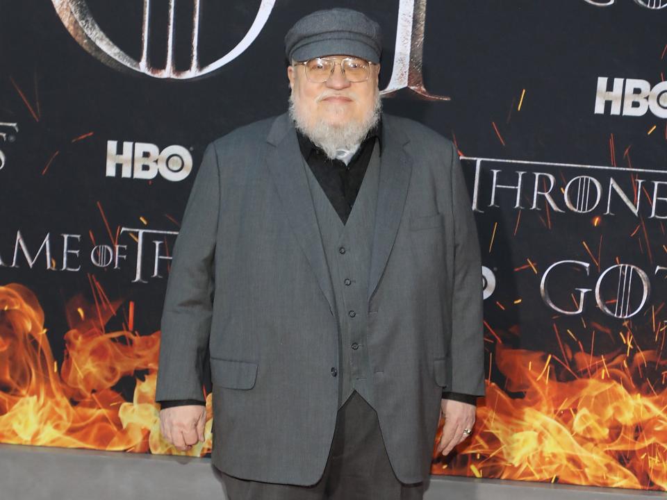 george r.r. martin at the season 8 game of thrones premiere, wearing a grey suit and cap and standing on the red carpet