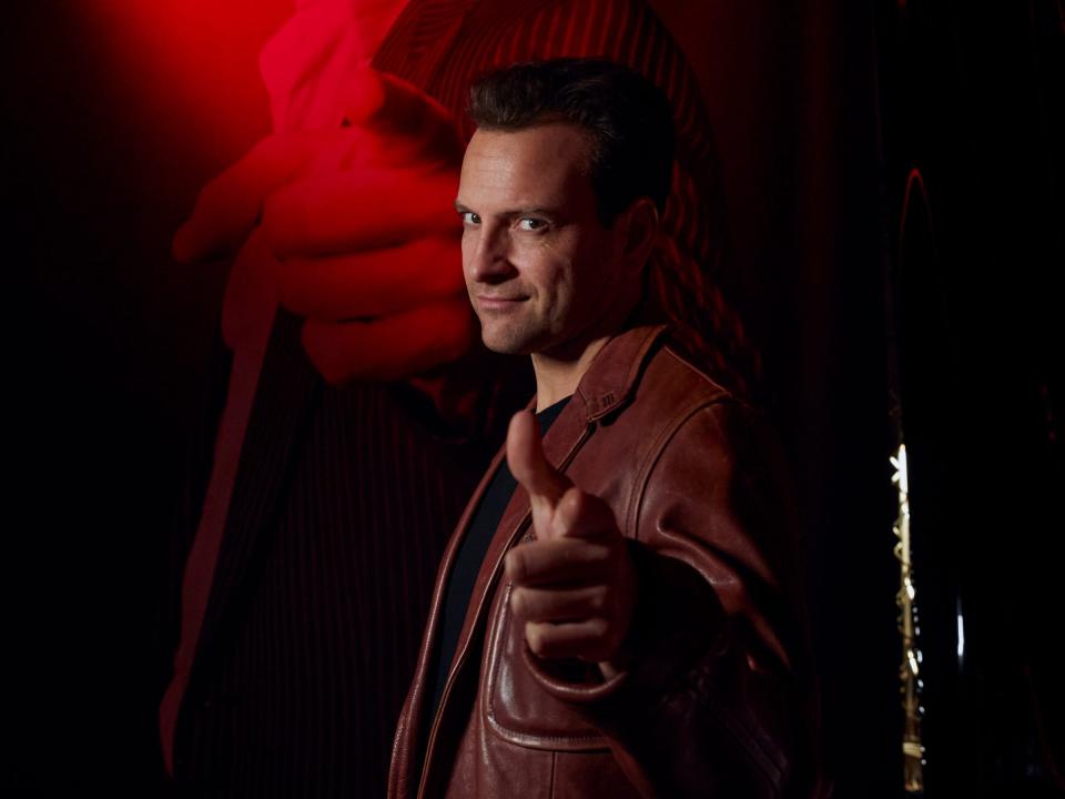 a man in a brown leather coat points a finger gun at the viewer, smirking
