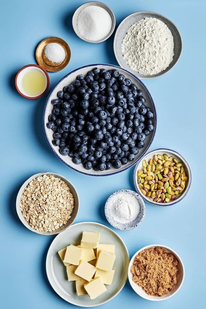 Ingredients for baking arranged neatly, including blueberries, flour, oats, sugar, butter, and pistachios