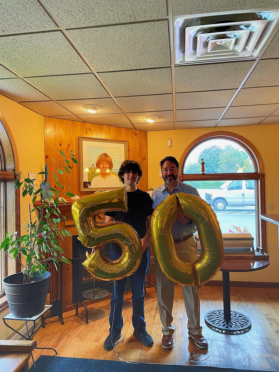 Symeon's Greek Restaurant owner Symeon Tsoupelis and his son, also named Symeon, pose with balloons commemorating the restaurants 50th anniversary, with a painting of the older Symeon's mother behind them.