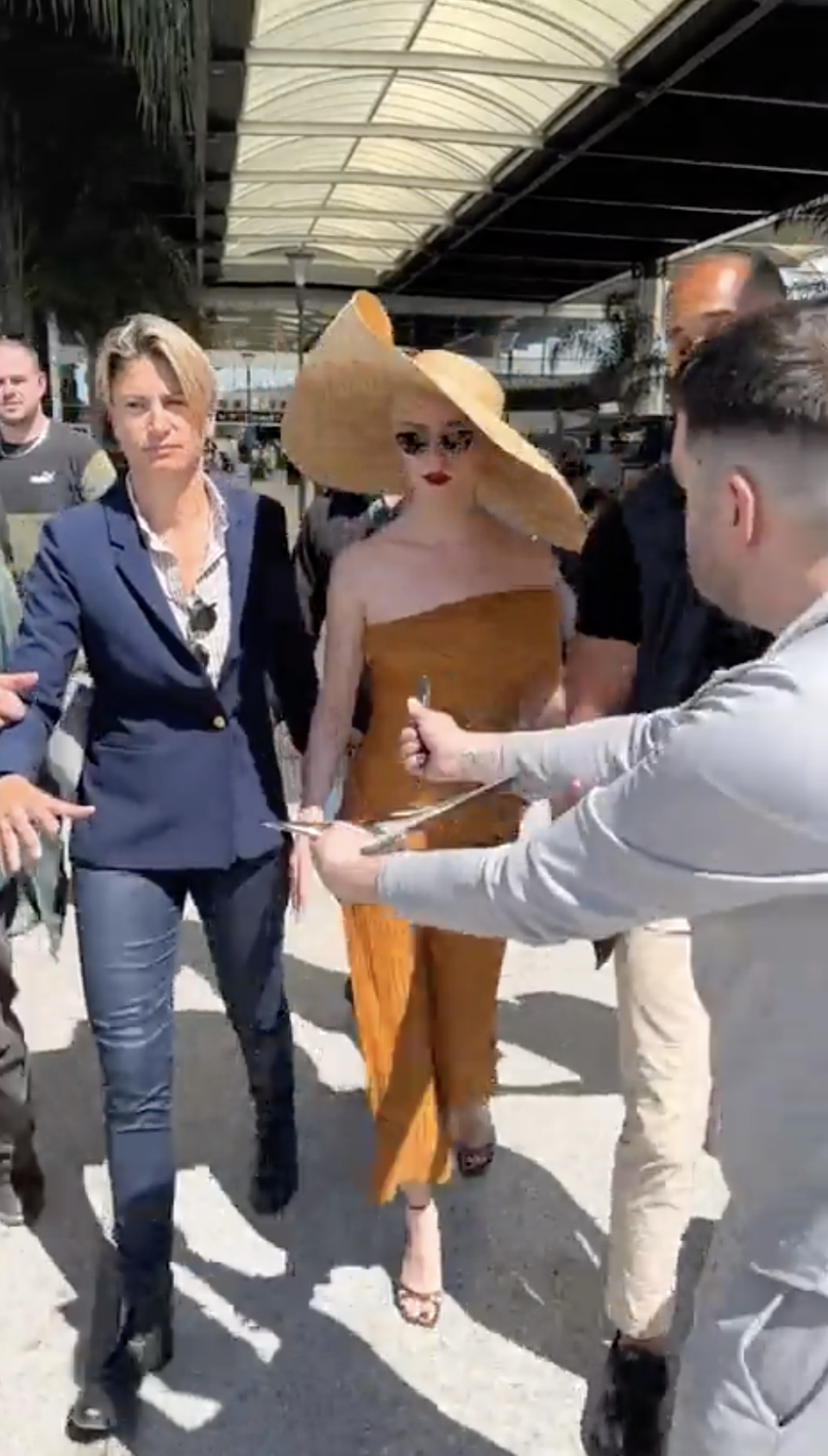 Anya Taylor-Joy is seen walking with several people. She is wearing a large wide-brimmed hat and an off-shoulder jumpsuit. Security and onlookers surround her