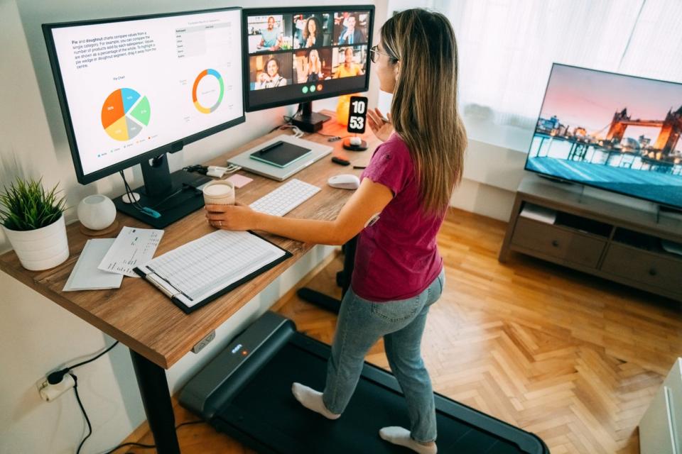 Woman in home office with two monitors, a walking pad desk, television, and phone. 