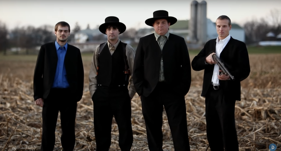 four men standing outside with one holding a rifle