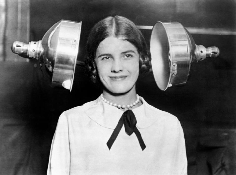 A smiling woman sits between two large metallic, round blowers that look as if they're about to squeeze the sides of her head