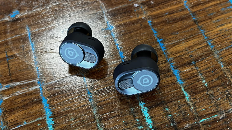 The Devialet Gemini II wireless earbuds shown on a textured wooden background