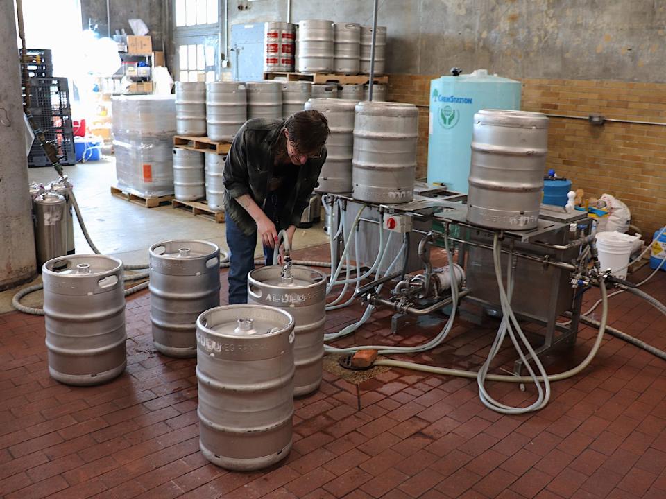 person in plaid flannel and jeans connects a tube to one of a dozen large metal canisters on a red brick floor