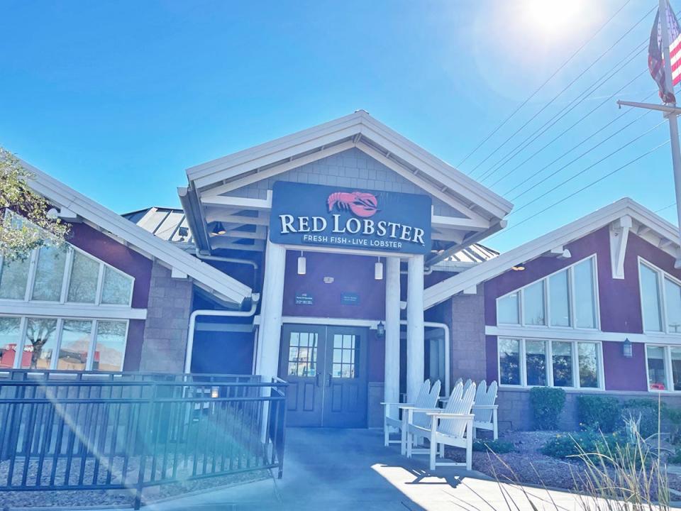 Exterior of Red Lobster. The building has peaked roofs and red siding and has white chairs by the entrance