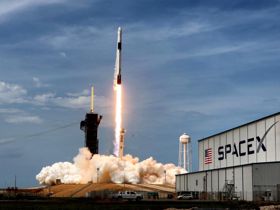 A SpaceX Falcon 9 rocket launches against a dusk sky, with a SpaceX branded factory visible in the foreground.