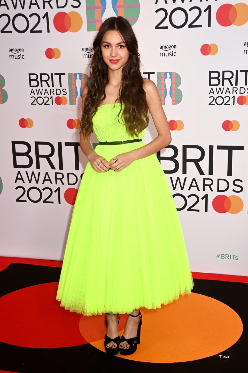 Olivia in a neon green/yellow strapless dress with a full tulle skirt that ends at her ankles. She has a black belt and black platform heels.