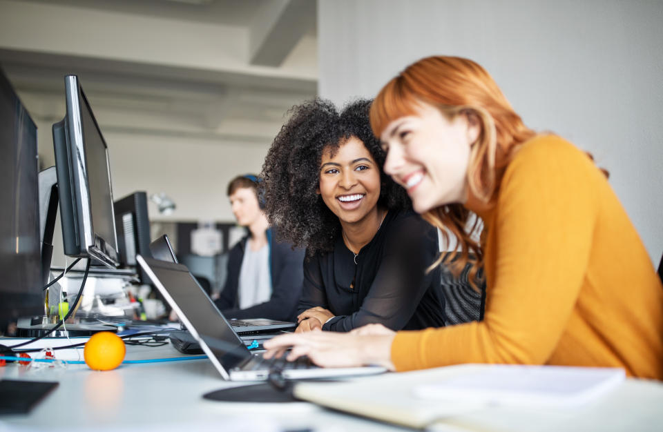 Two young women working together on laptop with male colleague in background. Two female colleagues in office working together.