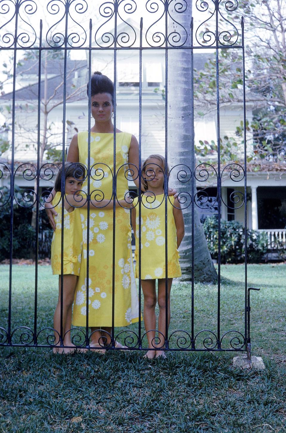 Lilly Pulitzer (center) stands with her daughters Liza (left) and Minnie outside of their Palm Beach home on South Lake Trail in 1963. Their yellow-patterned dresses with white trim have now become an iconic vintage look for which the Lilly Pulitzer brand is known.
