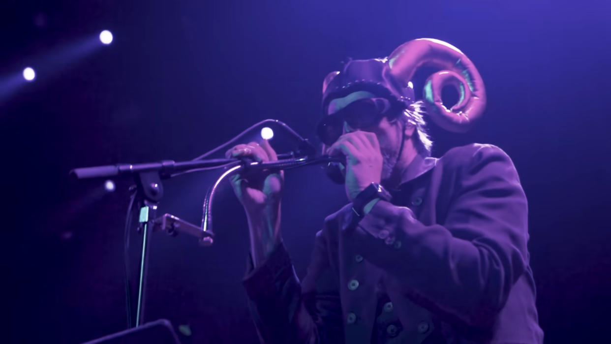  A picture of Les Claypool singing live on stage and wearing a ram-horned helmet 