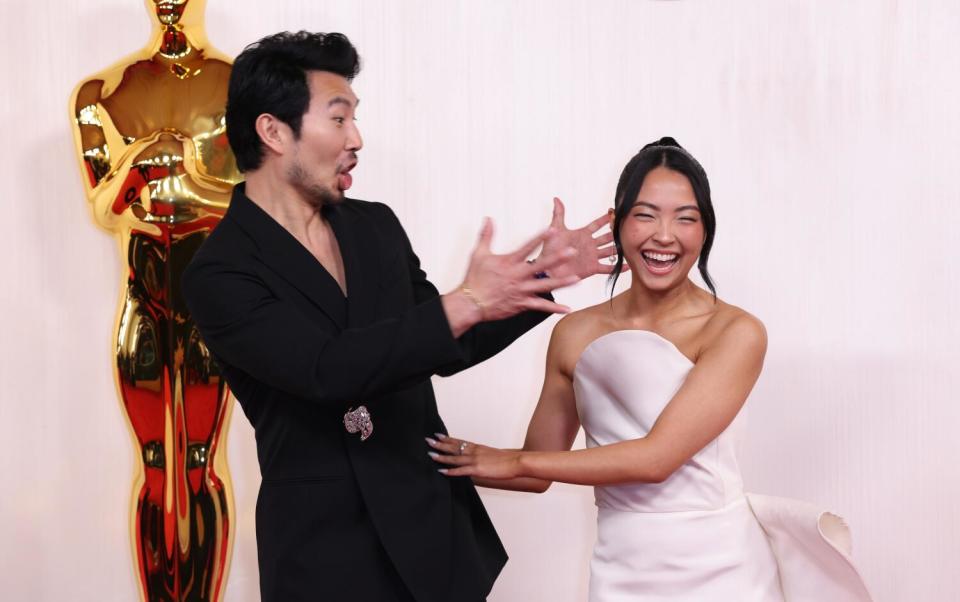 Simu Liu, in a black suit, and Allison Hsu, in a white gown, laugh in front of an Oscar statue.