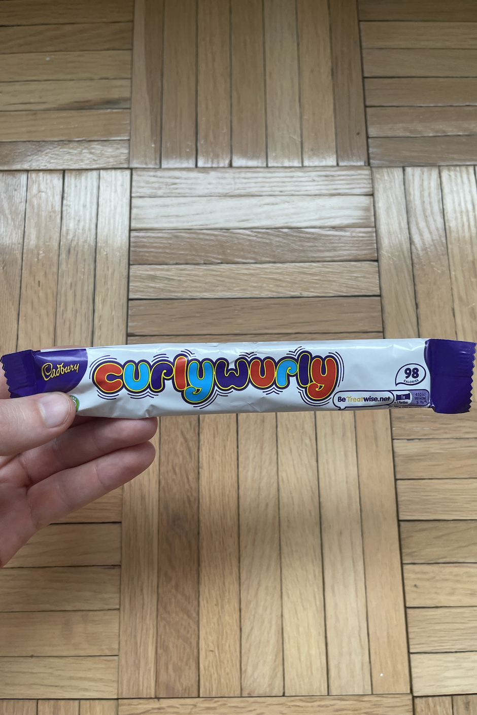 A person holding a Cadbury Curly Wurly chocolate bar against a wooden floor background