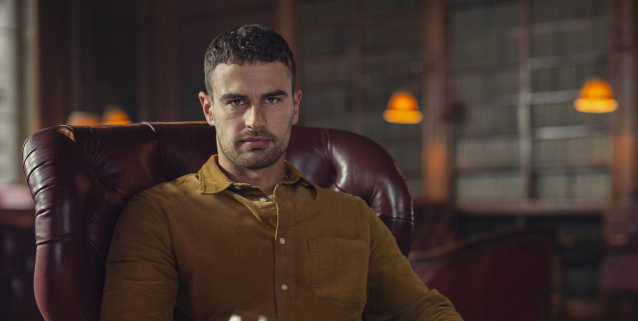 theo james in the gentlemen, a young man sits in an armchair looking at the camera and holding a wine glass