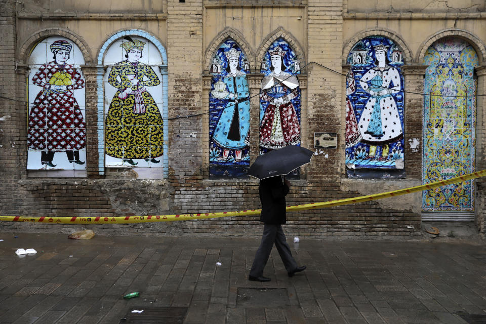 A man shelters from the rain with an umbrella a he walks past an old building decorated with replica of Iranian old paintings in a mostly empty street in a commercial district in downtown Tehran, Iran, Sunday, March 22, 2020. On Sunday, Iran imposed a two-week closure on major shopping malls and centers across the country to prevent spreading the new coronavirus. Pharmacies, supermarkets, groceries and bakeries will remain open. (AP Photo/Vahid Salemi)