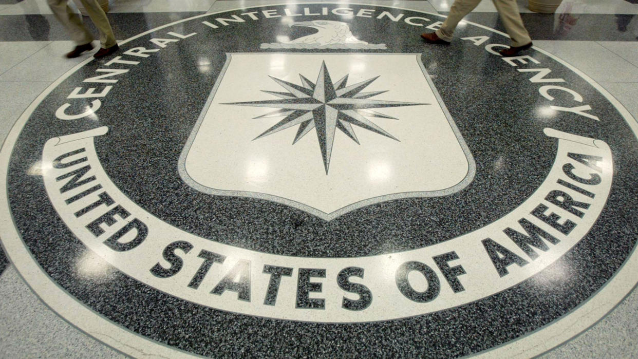 The CIA symbol, which reads Central Intelligence Agency United States of America, decorates a floor, with the legs of two people standing near its perimeter visible, both wearing khaki pants.