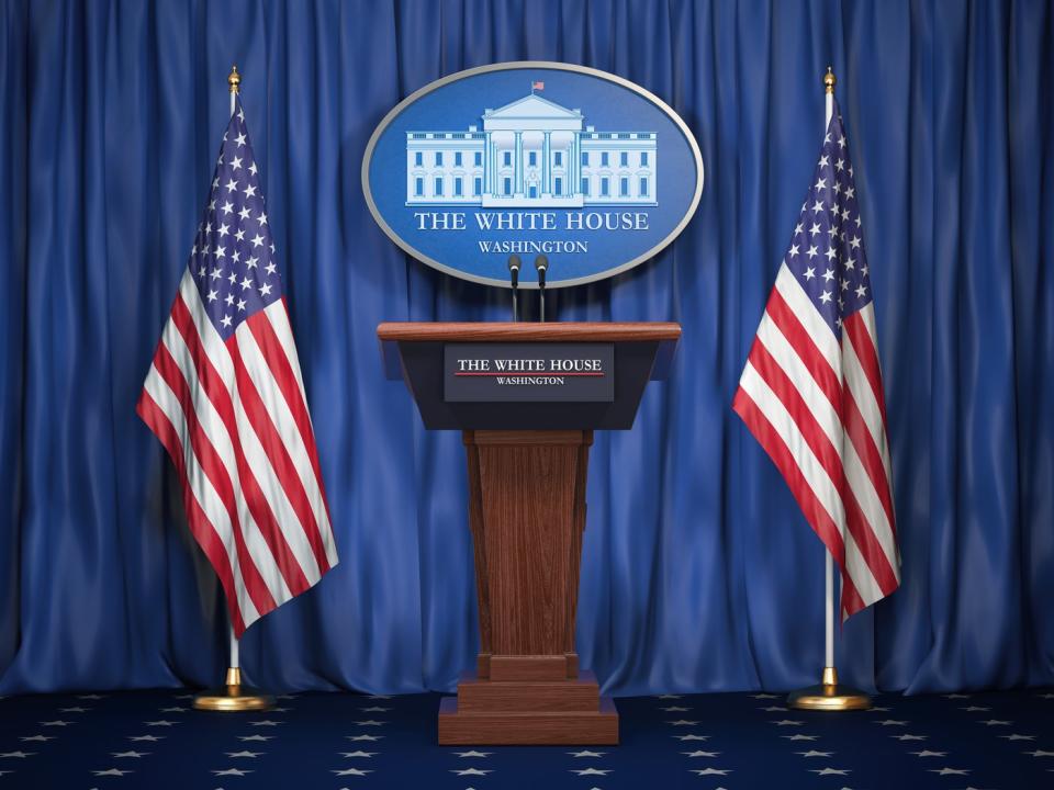 A podium between two US flags with the White House sign on the podium and on the curtains behind it.