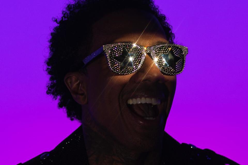 Backed by a glowing purple backdrop, Nick Cannon poses for a portrait in sparkly shades.