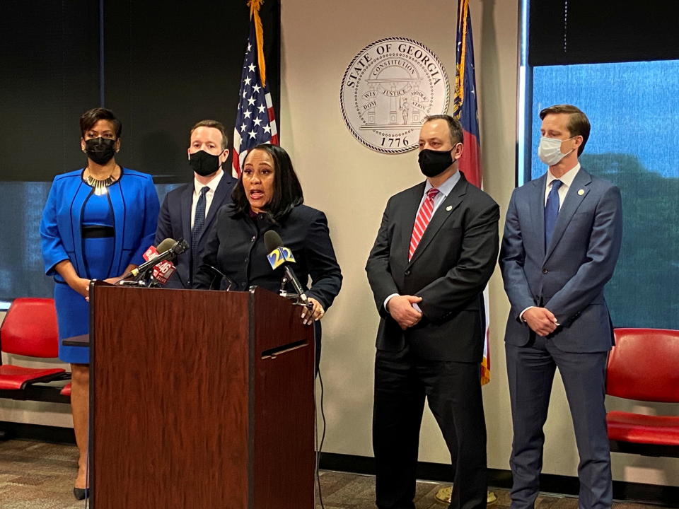 Fulton County District Attorney Fani Willis stands at a podium with microphones flanked by four people wearing face masks in front of the Georgia state seal.