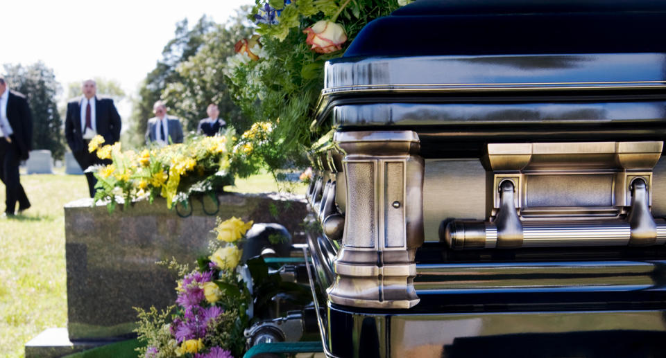 Funeral close-up of black casket with flowers near it and mourners visible in the background.