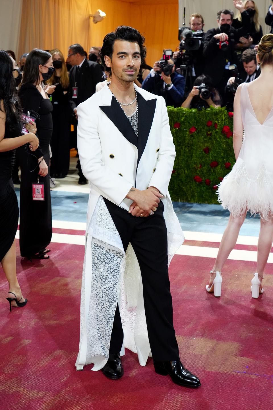 Joe in a white tuxedo jacket with black lapels and buttons and a white lace train cascading from the jacket. He's wearing a matching tank top underneath and black pants.