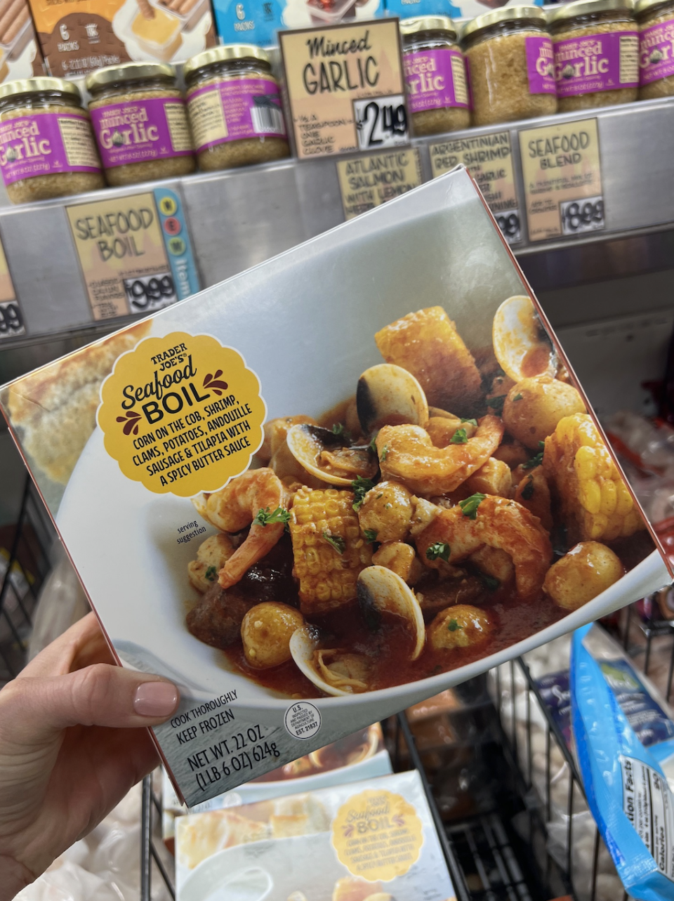 A hand holding a Trader Joe's Seafood Boil package with shrimp, corn, potatoes, andouille sausage, and seafood in a spicy butter sauce