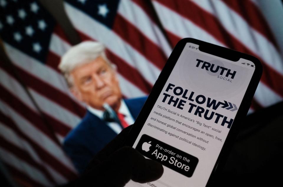A photo illustration showing a person checking the app store on a smartphone for "Truth Social", with a photo of former President Donald Trump in the background.