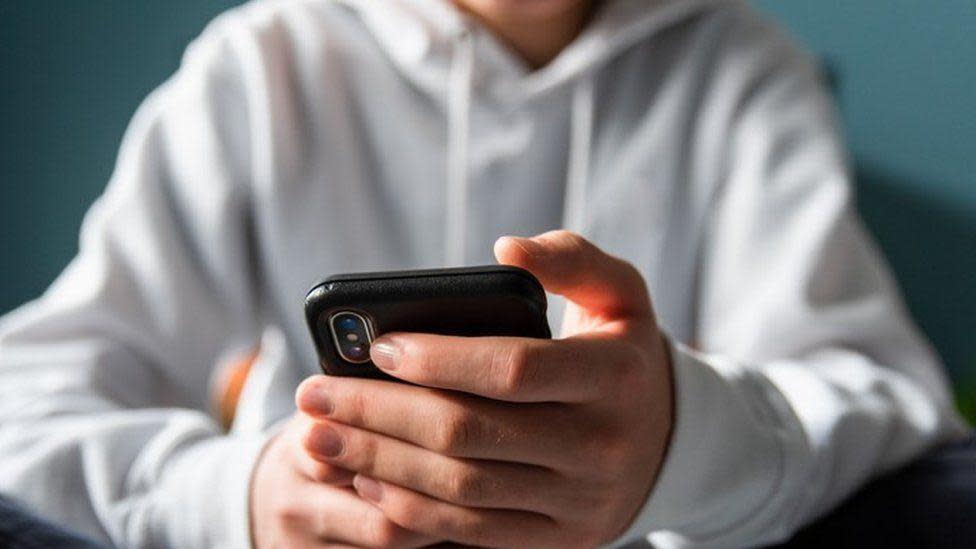 A boy, whose face is not visible and is wearing a white shirt, hold a smartphone in his hands