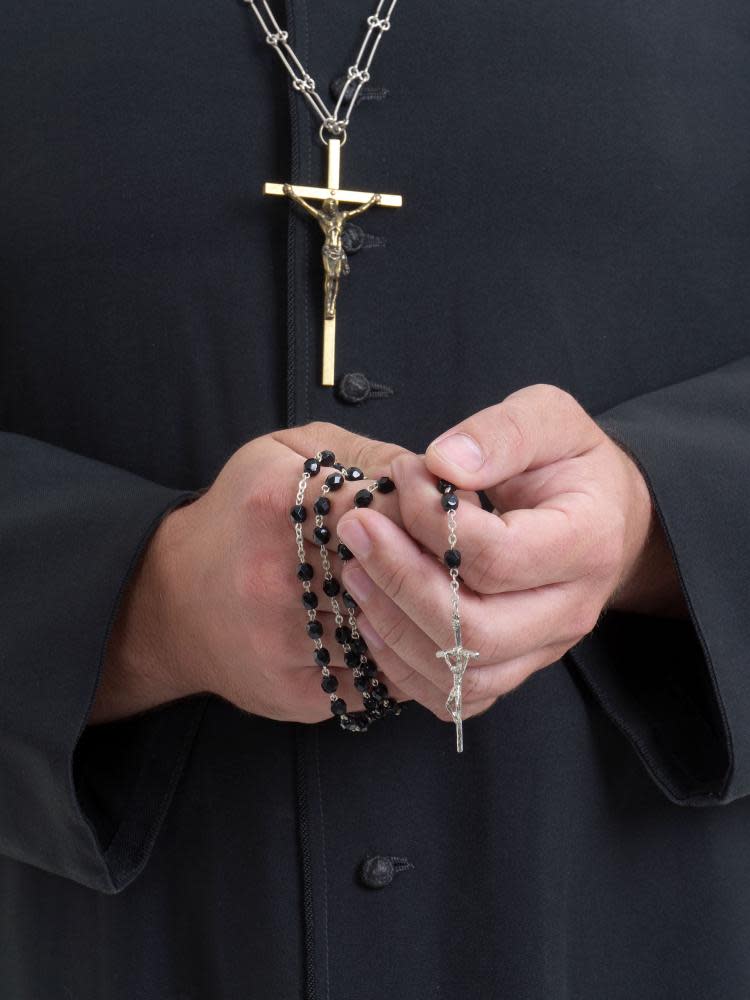 a figure wearing a black cassock with a gold crucifix holding black rosary beads in clasped hands