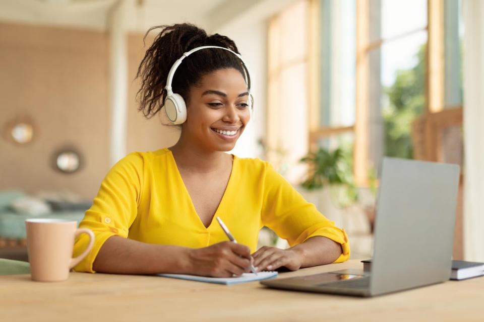 A smiling woman wearing headphones takes notes on a paper pad while looking at a computer screen.