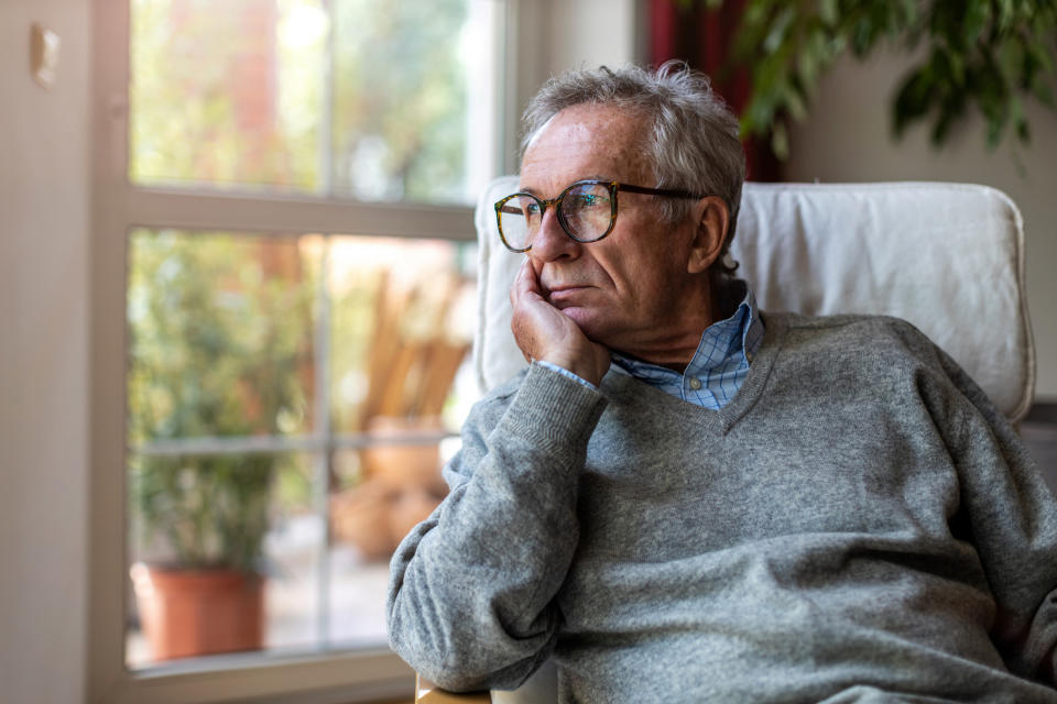 An older man with glasses and wearing a sweater looks out the window thoughtfully, resting his head on his hand