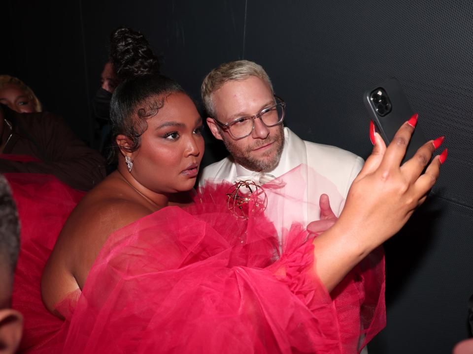 lizzo, with red nails and a poofy red dress, takes a selfie with seth rogen, wearing a white suit, while holding her emmy award