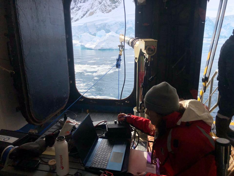 A woman inside a cruise ship with a laptop and other equipment and the Antarctic landscape outside the window