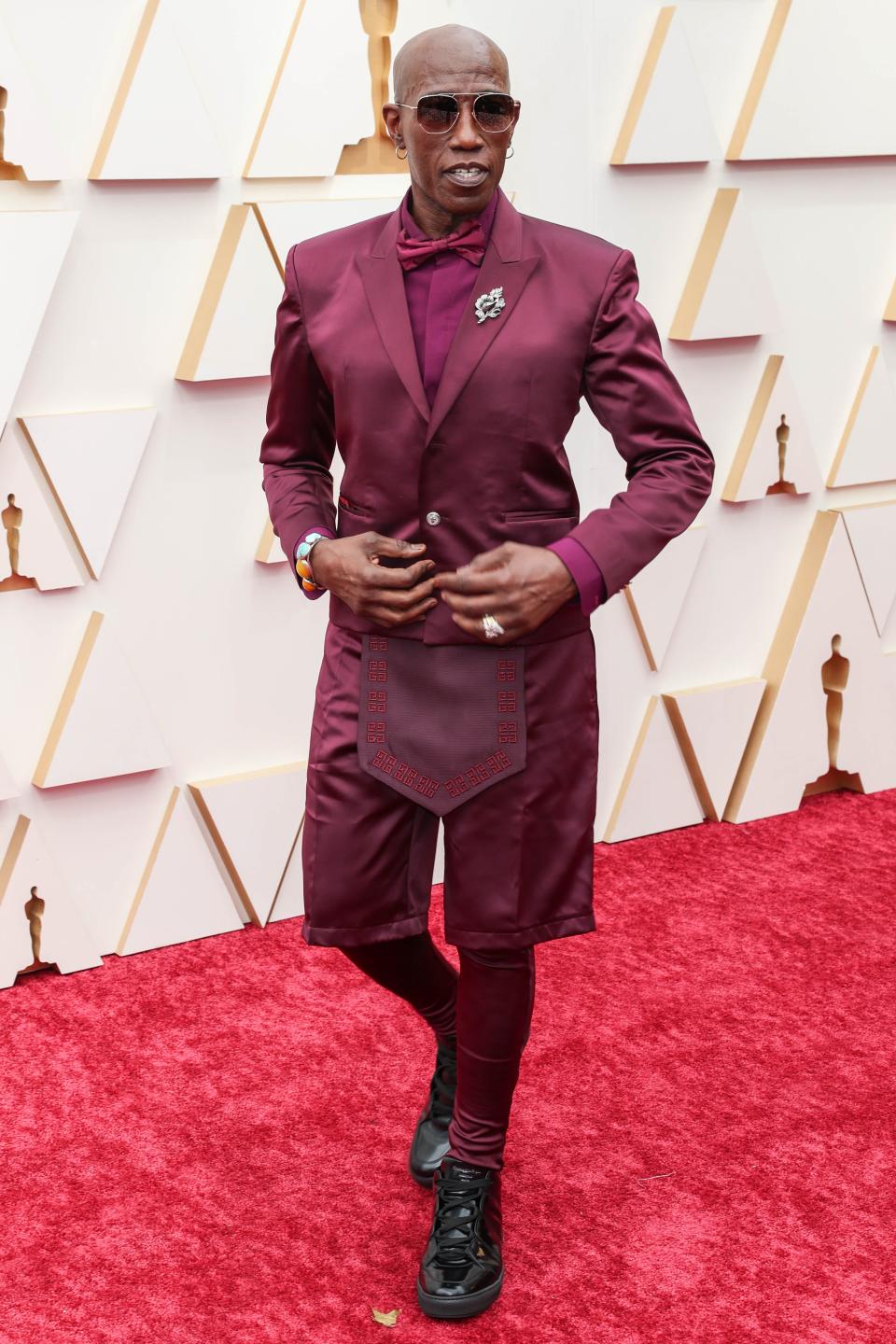 Wesley in a satin burgundy suit jacket, shirt, bow tie, and shorts over legging pants. There's a panel over the shorts. He's wearing sunglasses and black chunky sneaker shows.