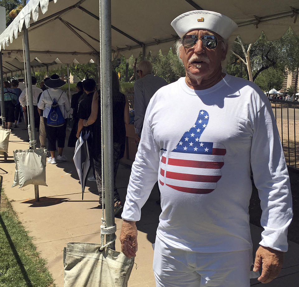 Louis Albin, an artist in Phoenix and Navy veteran, displays his custom-made T-shirt, as he waited in line for the public viewing of the late Sen. John McCain Wednesday, Aug. 29, 2018, outside the Arizona Capitol. The shirt, which features an image of a thumb-up covered in the stars and stripes of the flag, is a play on McCain's thumb-down vote that sank President Donald Trump's repeal of the Affordable Care Act. Albin turned the thumb up as a wink to McCain's vote, which Albin approved of. (AP Photo/Jacques Billeaud)