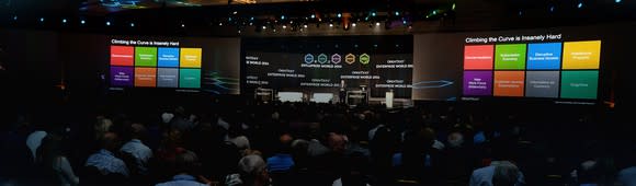 OpenText conference presentation, with speaker in front of large audience and colored displays showing a corporate presentation.