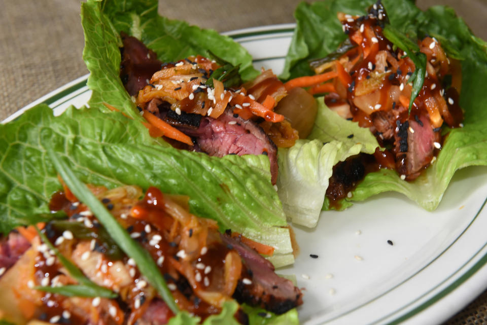 Three lettuce wraps filled with sliced beef, shredded carrots, green onions, and drizzled with a sauce, garnished with sesame seeds on a white plate