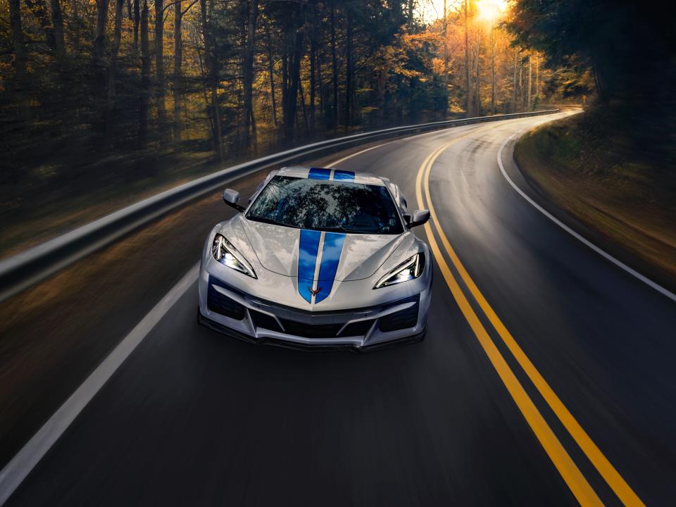 A white sports car with blue stripes drives down a road with trees in the background.