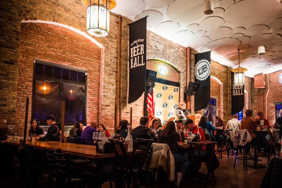 People at long tables at an indoor bar, with stained-glass lights, brick walls, and signs that read "Lakefront Brewery Beer Hall." 