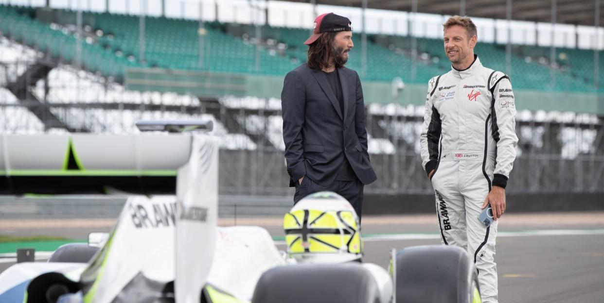 keanu reeves and jenson button talk to each other and smile as they stand behind a brawn formula 1 car on a racing circuit