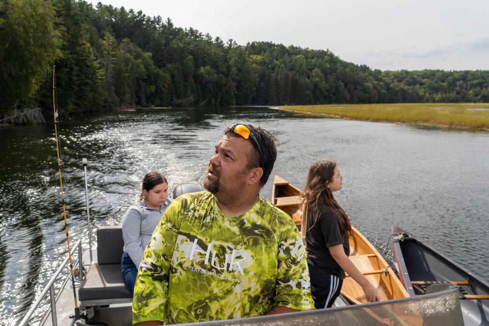 Bay Mills Community College farm technician Dennis Carrick looks above pine trees toward the sounds of eagles overlooking the water while driving a boat towing canoes to a section of the Au Sable River in Oscoda on Saturday, Sept. 16, 2023, before harvesting manoomin "wild rice" growing on the water with Jasmine Cabarrubia, left, of Manistique, and River Bailey, of Brimley. "They're watching us and guiding us," said Carrick, of the birds sacred to Native Americans.