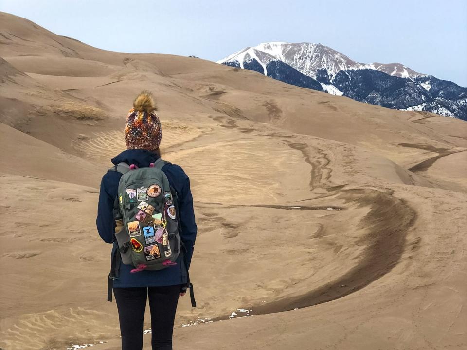 Emily, wearing a backpack covered in patches, a black jacket, black leggings, boots, and a winter hat, walks toward large sand dunes and mountains.