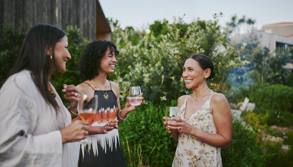 Three women enjoying drinks and laughing together in an outdoor garden setting. The woman on the right wears a light floral dress, and others wear casual attire. Names unknown