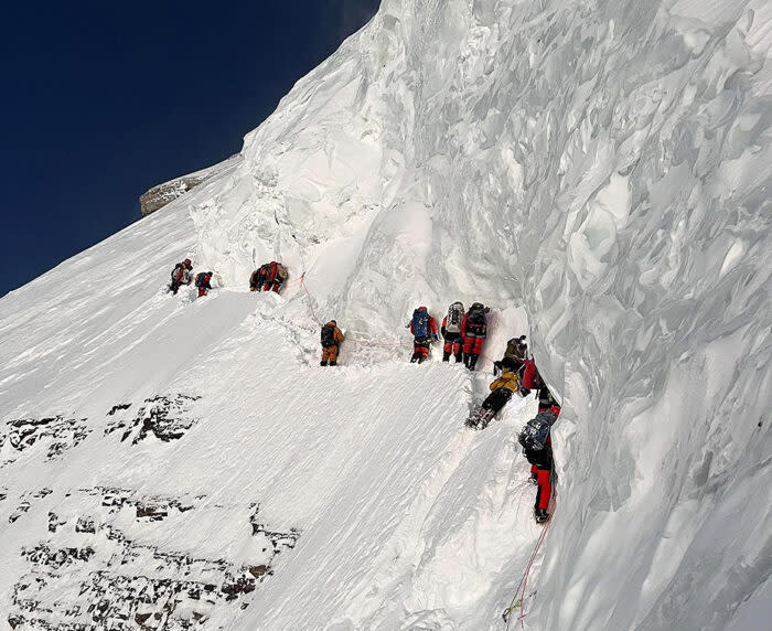 a line of climbers, attached to a rope, crosses a snow slope below a giant serac.