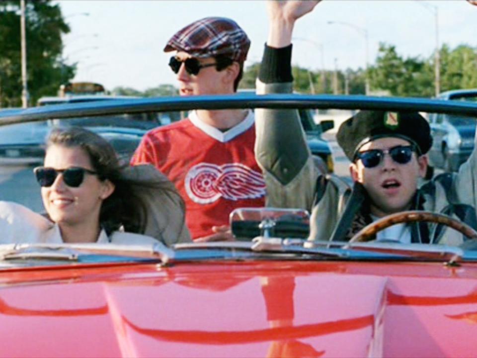 Mia, Alan, and Matthew sitting in an open-top red Cadillac all in sunglasses and with Matthew's arms in the air.