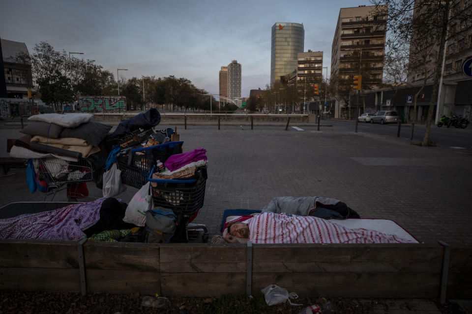 In this Friday, March 20, 2020 photo, Boris, 42, from Bulgaria, sleeps under a blanket in the street in Barcelona, Spain. While Spanish authorities tell the public that staying home is the best way to beat the coronavirus pandemic, some people are staying out because home has come to mean the streets of Madrid and Barcelona. Authorities are scrambling to get as many homeless people off the streets without cramming them into a group shelter, where the risk of getting infected with the virus could be even greater. (AP Photo/Emilio Morenatti)