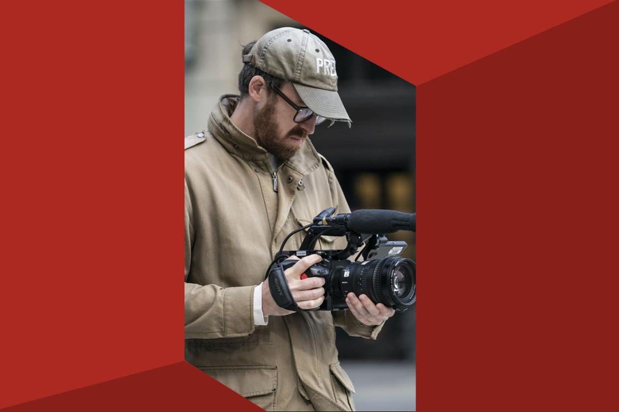 A white man in a baseball cap holds a camera on a New York street.