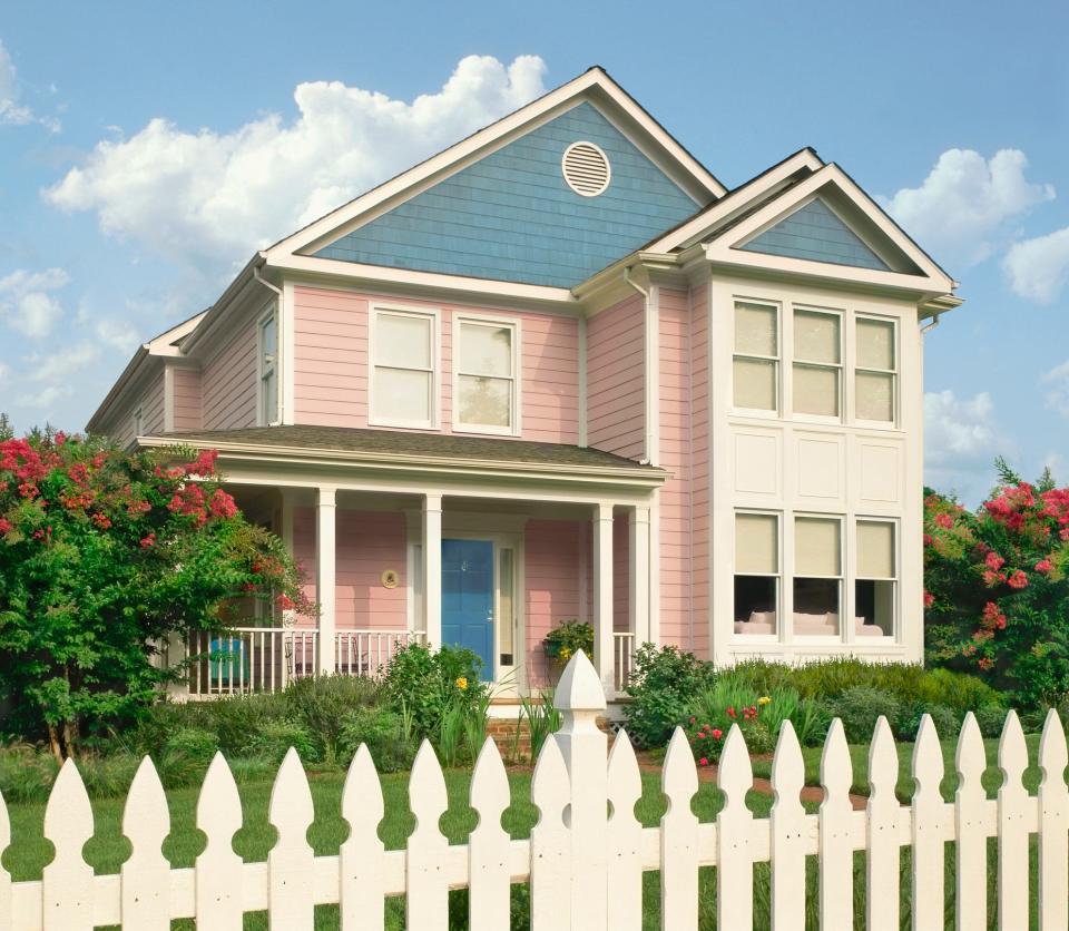 A two story house with pale pink and pale blue siding is shown. A white fence is in the foreground.