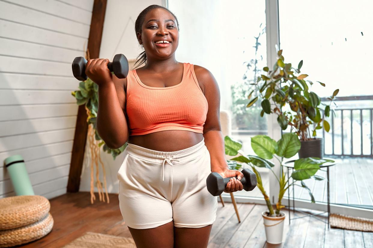 Positive african american woman using dumbbells for sport training at cozy modern house. Overweight young lady wearing orange sport bra and shorts strengthening arm muscles.